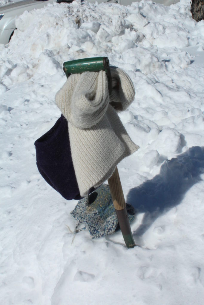 hat, mittens, gloves and scarf hanging from an unused shovel in a pile of snow.